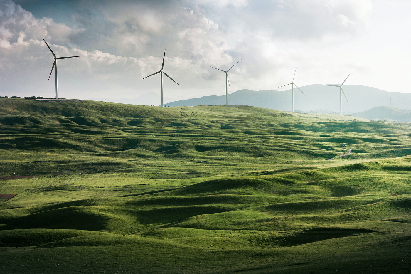 Photograph of wind turbines sitting on rolling, green hills. A blue sky and clouds are in the background.